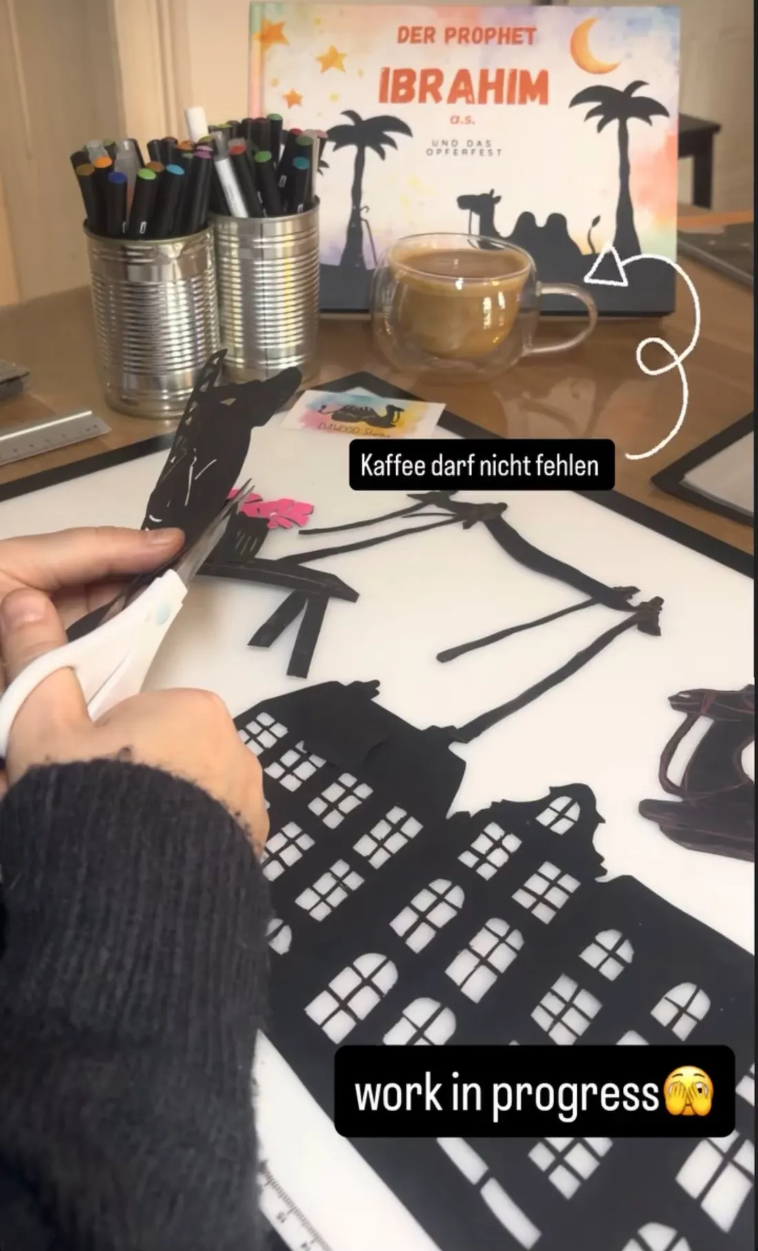 Antje schneidet Schattenfiguren von Kamel und Stadt aus schwarzem Papier, daneben Kaffee und Stifte